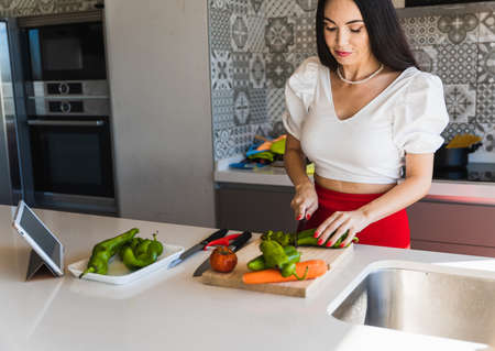 Woman in stylish outfit cutting fresh pepper near tablet with online recipe while preparing vegetarian salad for lunch at homeの写真素材