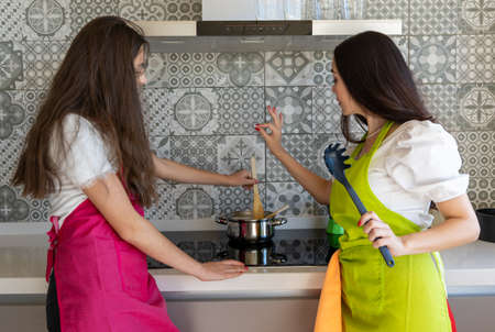 Mother adding salt into dish while daughter stirring food in saucepan while preparing lunch in contemporary kitchen at homeの写真素材