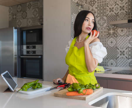 Housewife in green apron holding ripe tomato and looking away while cutting vegetables for healthy salad near tablet in kitchen at homeの写真素材