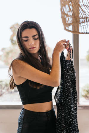 Young brunette in casual clothes holding trendy dress while getting dressed against window on sunny day at homeの写真素材