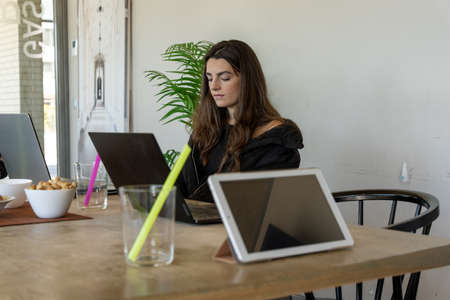 Young woman with long hair sitting at table near digital devices and snacks and working on remote project in cafeteriaの写真素材