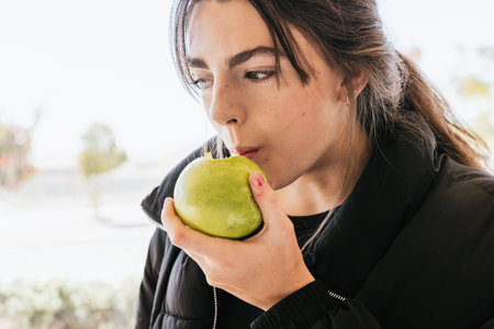 Young female in outerwear biting ripe apple while having healthy lunch on blurred background on sunny dayの写真素材