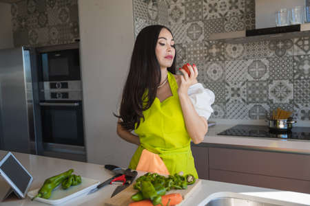Brunette in apron holding hand on waist and inspecting ripe apron during healthy dish preparation in contemporary kitchen at homeの写真素材