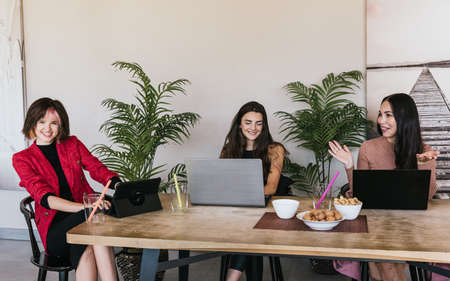 Gleeful stylish women in casual clothes using digital devices and smiling while sitting at cafeteria table and talking during remote jobの写真素材