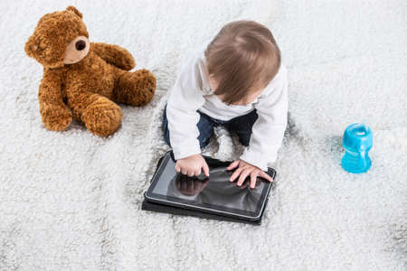 Stock studio photo with a white background of a baby touching the screen of a tablet with a teddy bear on the side and a container with handles to drink.の写真素材