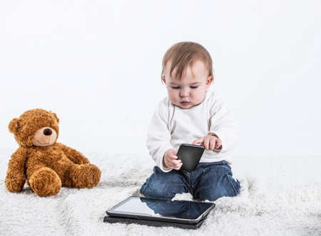 Stock studio photo with a white background of a baby touching the screen of a tablet with a teddy bear on the side and a container with handles to drink.の写真素材