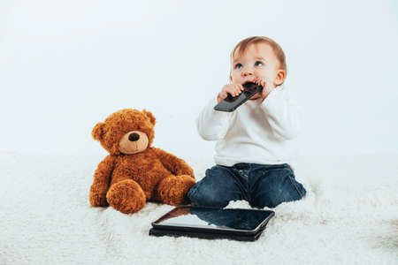 Stock studio photo with the white background of a baby's face with a mobile in his mouth next to a teddy bear and a tablet.の写真素材