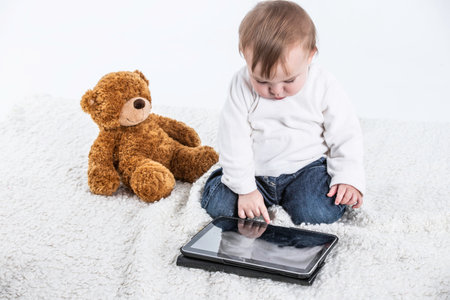 Stock studio photo with a white background of a baby touching the screen of a tablet with a teddy bear on the side and a container with handles to drink.の写真素材
