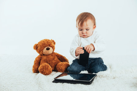Stock studio photo with a white background of a baby looking at the screen of a tablet with a teddy bear next to it.の写真素材
