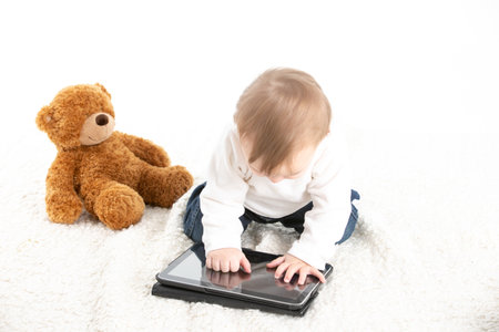 Stock studio photo with a white background of a baby touching the screen of a tablet with a teddy bear next to itの写真素材