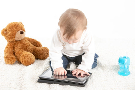 Stock studio photo with a white background of a baby touching the screen of a tablet with a teddy bear on the side and a container with handles to drink.の写真素材