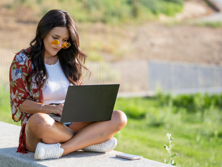 Young woman in sunglasses using a laptop computer while sitting on a bench in a park. Technology concept.の写真素材