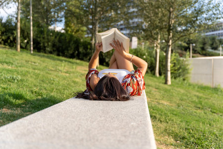 Woman with sunglasses enjoying reading a book while relaxing lying down on a bench in a park.の写真素材