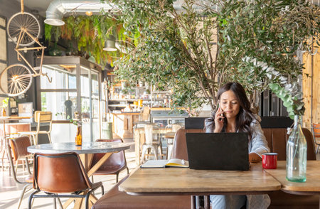 Woman talking on the phone while working online with a laptop computer in a coffee shop. Technology and business concept.の写真素材