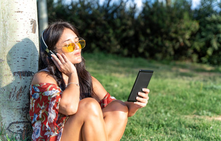 Woman with sunglasses listening music with headphones while reading an ebook in a park. Technology concept.の写真素材