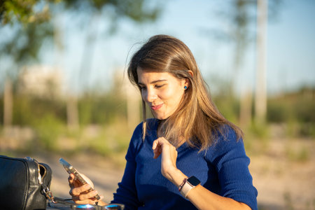 Woman using her mobile phone and smiling while sitting on a table outdoors in the nature. Technology concept.の写真素材