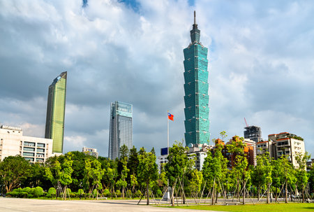 A tall green skyscraper dominates the skyline of Taipei, Taiwan, surrounded by contemporary high-rises, lush trees, and the national flag, set against a backdrop of dramatic clouds.の写真素材