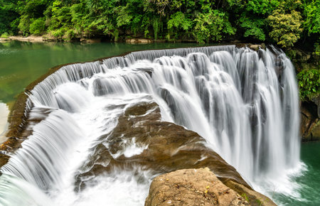 Shifen Waterfall in Pingxi District of New Taipei, Taiwan, cascades powerfully into a turquoise pool amid dense green foliage, offering a scenic and tranquil natural attraction often called the Little Niagara of Taiwanの写真素材