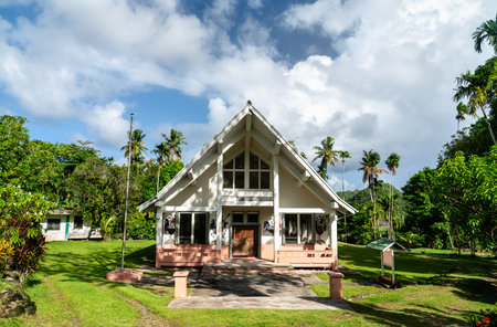 Traditional Palauan meeting house Bai ra Mlengl, nestled in lush tropical surroundings in Airai. The building features a steep gabled roof and distinctive painted motifs, standing peacefully amid palm trees and vibrant greenery under a partly cloudy skyの写真素材