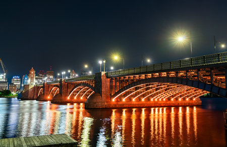 Night view of the Longfellow Bridge in Boston glowing with orange lighting, arching over the Charles River with reflections shimmering on the water and the city skyline in the backgroundの写真素材