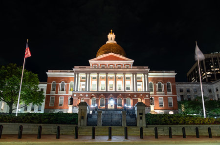 Night view of the Massachusetts State House in Boston with its iconic golden dome lit against the dark sky, flanked by flags and classical architectureの写真素材