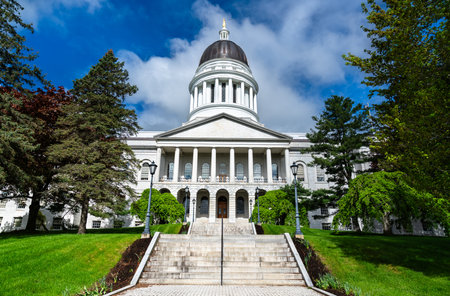 Classical white granite capitol building with a prominent dome and columned portico, surrounded by manicured green lawns and tall trees in Augusta, Maine, USAの写真素材