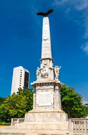 Marble obelisk topped with an eagle stands prominently in Centenario Park in Cartagena, Colombia. Classical statues adorn the base, and lush greenery surrounds the monument against a clear blue skyの写真素材