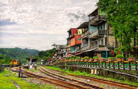 Colorful hillside houses overlook the curving railway tracks and a small diesel locomotive at Shifen Station on the Pingxi line in New Taipei, Taiwan, with lush forested hills beneath a cloudy summer skyの写真素材