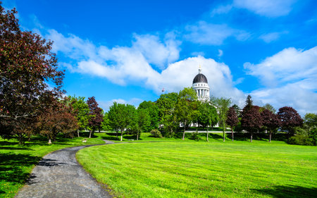 In Augusta, United States, the Maine State House dome peeks above leafy Capitol Park, where a curved gravel path winds across bright spring lawns under a vivid blue sky.の写真素材