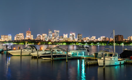 At night in Cambridge, United States, small yachts rest in a Charles River marina while Boston illuminated skyline glows across the water, reflections shimmering under a clear blue-black sky.の写真素材