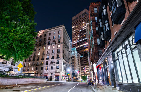Night scene in Boston, Massachusetts, United States, shows a quiet downtown street lined by historic brick buildings and a modern high rise, tree foliage glowing under streetlights and dark sky.の写真素材