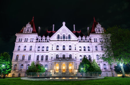 Albany, United States, the ornate New York State Capitol glows white and red against a dark sky, its steep mansard roof and arched windows mirrored in a quiet plaza fountain.の写真素材