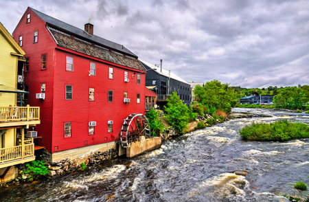 In Littleton, New Hampshire, a red clapboard grist mill with a working waterwheel stands above the rushing Ammonoosuc River beneath a bright, cloud-dappled summer sky.の写真素材