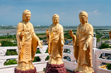 Three gold Buddha statues stand on a terrace at Fo Guang Shan Monastery in Kaohsiung, Taiwan, raised above the plains and river under a clear summer skyの写真素材