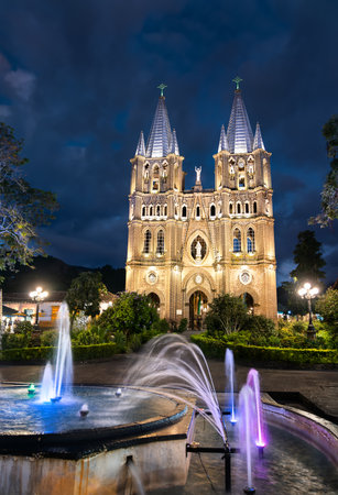 In Jardin, Colombia, the twin-spired Basilica of the Immaculate Conception glows gold at dusk as color-lit fountains arc in the leafy plaza before it.の写真素材