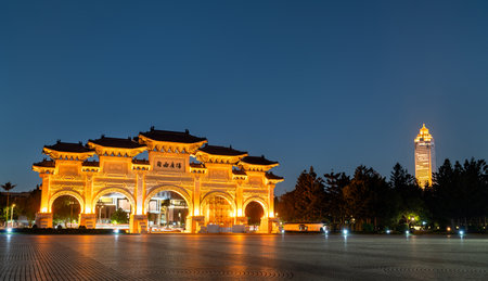 Wide twilight view of the Liberty Square Arch glowing with golden flood-lighting, its ornate traditional roof silhouettes mirrored by a distant tower beyond the spacious plazaの写真素材