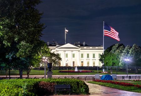 The White House, the official residence of the U.S. President, viewed from Lafayette Square in Washington, D.C. The historic neoclassical building with a United States flag are illuminated at night behind a security fenceの写真素材