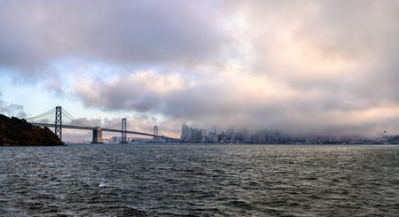 A moody, panoramic view of the San Francisco skyline mostly hidden by thick, low fog. The Bay Bridge is visible on the left, spanning the choppy bay water under a dramatic, cloudy skyの写真素材