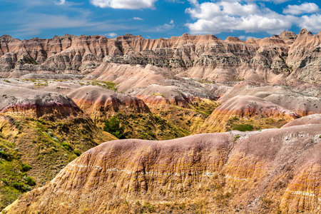 Colorful sedimentary rock layers in Badlands National Park, South Dakota. Eroded yellow and red hills rise under a blue sky with cloudsの写真素材