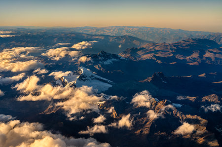 Aerial view of the Andes Mountains in Peru. Golden sunrise light illuminates the high peaks, while deep valleys and a sea of clouds are in shadowの写真素材
