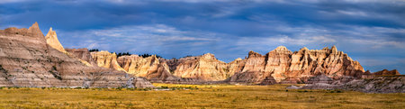 Panoramic view of eroded rock formations in Badlands National Park, South Dakota. Rugged buttes rise from the grassy prairie under a dramatic blue sky with dark cloudsの写真素材