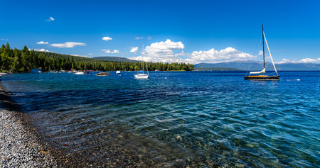 View of Lake Tahoe from the shore near Tahoe City, California. Boats and a sailboat float on the clear blue water with mountains in the background under a sunny skyの写真素材