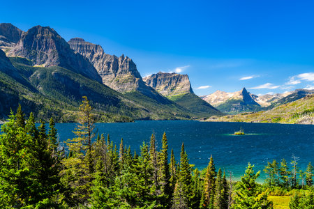 Panoramic view of the vibrant blue Saint Mary Lake, featuring Wild Goose Island, in Glacier National Park. The Rocky Mountains rise grandly under a clear blue sky, framed by pine treesの写真素材