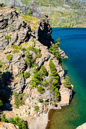 Rocky cliffs rise above blue waters of Saint Mary Lake at Sun Point in Glacier National Park Montana. Landscape features burnt tree snags from past wildfires standing on rugged promontoryの写真素材
