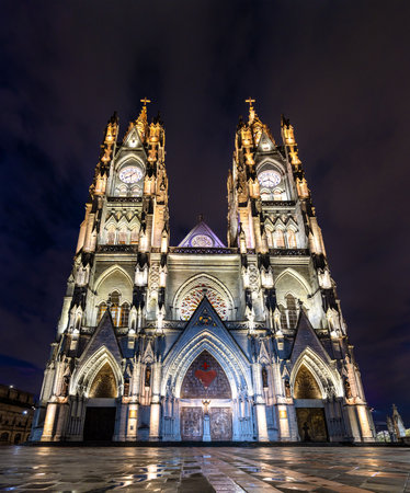 Illuminated facade of Basilica del Voto Nacional rises above wet plaza in Quito, Ecuador. Neo-Gothic church features twin towers and intricate stone carvings under dark night skyの写真素材