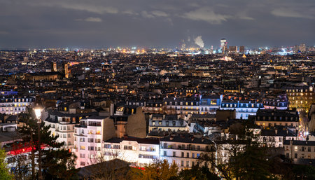 Cityscape of Paris, France glows at night seen from Montmartre hill. Panoramic view features illuminated rooftops, streetlights, and urban horizon under cloudy sky in Ile-de-France regionの写真素材