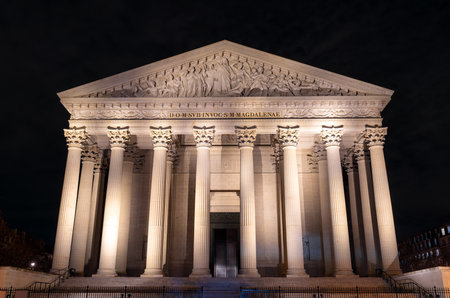 Illuminated La Madeleine church stands with Corinthian columns in Paris, France. Neoclassical Catholic temple features majestic stone facade and pediment sculpture under night skyの写真素材