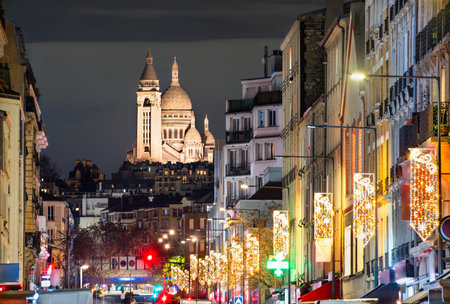 Illuminated Sacre-Coeur Basilica rises above city street at night in Paris, France. View from Saint-Ouen features festive Christmas lights and apartment buildingsの写真素材