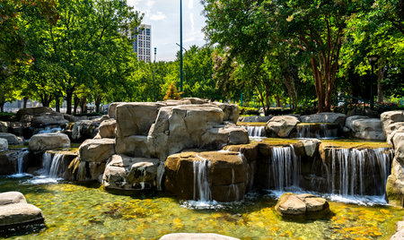 Centennial Olympic Park in Atlanta, Georgia. Scenic man-made waterfall flows over rock formations surrounded by green trees and city buildings under a blue skyの写真素材