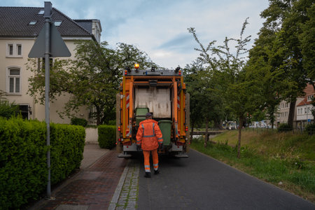 An orange garbage truck stands on a narrow green street, surrounded by green bushes and trees, a garbage man in uniform loads green garbage cans into the car. rear view. selective focus. Blurred backgroundの写真素材